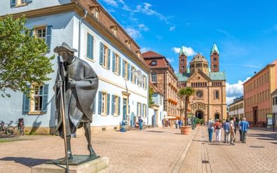 Bronzen standbeeld van een pelgrimsfiguur staat voor de Dom van Speyer met zijn kenmerkende tweelingtorens. Toeristen lopen over het geplaveide plein.
