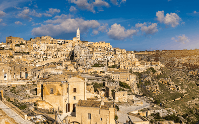 Panoramisch uitzicht op Matera's oude stenen gebouwen uitgehouwen in rotsachtige helling onder blauwe hemel met witte wolken.
