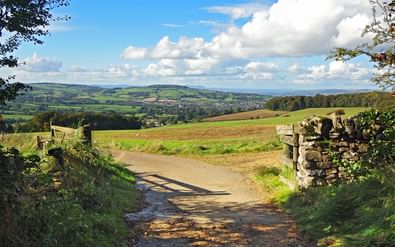 Landelijk pad met stenen muren door glooiende groene velden en heuvels in de Cotswolds onder bewolkte blauwe hemel.