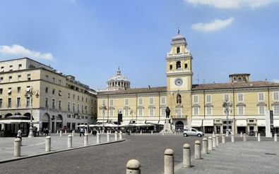 Piazza Garibaldi in Parma met gele barokgebouwen, klokkentoren, koepel en stenen paaltjes onder blauwe hemel met witte wolken.
