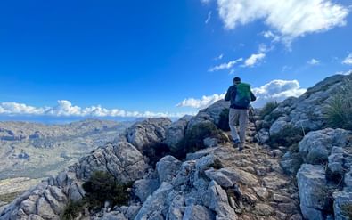 Wandelaar met groene rugzak op rotsachtig stenen pad door kalksteenterrein in Mallorca bergen onder blauwe hemel met wolken.