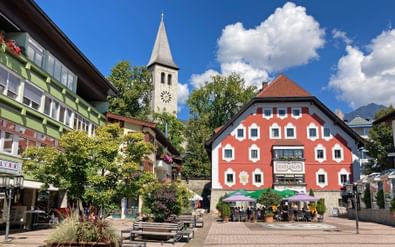 Kleurrijke gebouwen in het centrum van Saalfelden met witte kerktoren, café-terras en bergen op de achtergrond onder blauwe lucht.