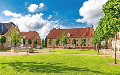 Groen gazonplein in Christiansfeld met gele bakstenen gebouwen met rode pannendaken. Jonge bomen staan langs het plein onder een blauwe lucht.