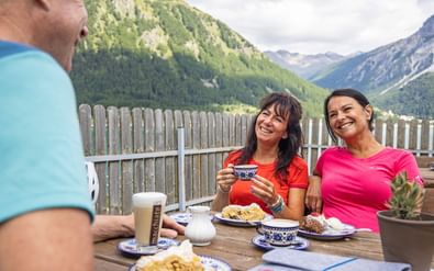 Drie fietsers genieten van koffie en taart aan buitentafel met bergdaluitzicht. Twee vrouwen in kleurrijke shirts glimlachen naar camera.