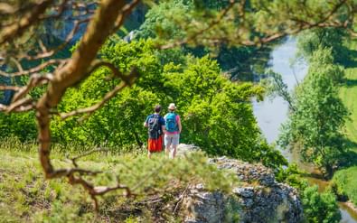 Twee wandelaars op rotsachtige klif bij Zwölf Apostel in Altmühltal, uitkijkend over een kronkelende rivier omgeven door groene bomen.