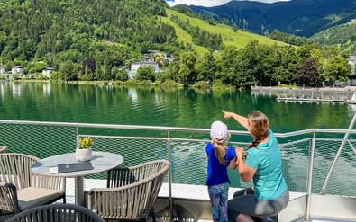 Moeder en kind op een bootterras bij Zeller See, wijzend naar groene bergen. Tuinmeubilair zichtbaar, omringd door helder water en alpenlandschap.