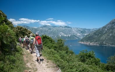 Groep wandelaars op een bergpad in Montenegro met uitzicht op een blauwe baai omringd door bergen en groene vegetatie.