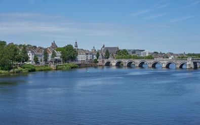 Stenen brug met meerdere bogen over een brede rivier in Maastricht. Historische gebouwen en kerktorens langs de waterkant onder blauwe lucht.