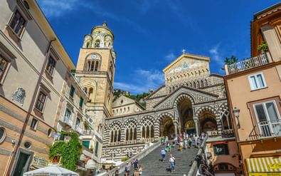 Kathedraal van Amalfi met gestreepte gevel en klokkentoren, stenen trappen naar de ingang, omringd door kleurrijke Italiaanse gebouwen.