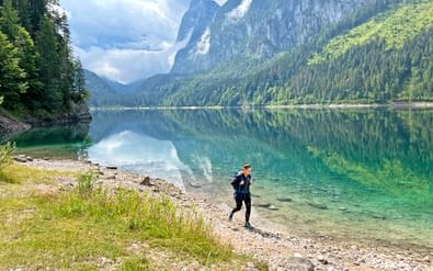Vrouwelijke wandelaar loopt langs de oever van het Gosausee meer met kristalhelder turquoise water dat dramatische Dachstein bergtoppen weerkaatst.
