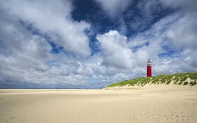 Rood-witte vuurtoren staat op met gras begroeide zandduinen met uitzicht op een breed zandstrand op Texel onder dramatische bewolkte hemel.