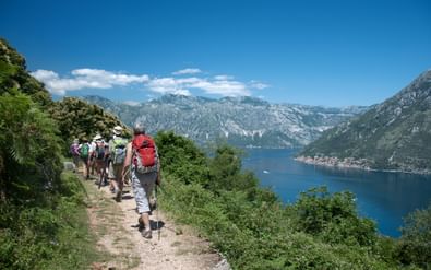 Groep wandelaars op een bergpad in Montenegro met uitzicht op een blauwe baai omringd door bergen en groene vegetatie onder heldere hemel.