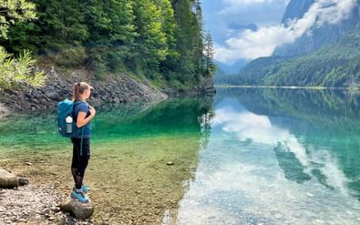 Vrouwelijke wandelaar met rugzak staat op rotsachtige oever van kristalhelder Gosausee, omringd door beboste bergen en dramatische pieken.