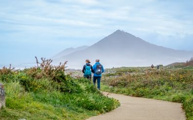 Twee wandelaars in blauwe jassen op kustpad bij Moledo met bergtop in mistige achtergrond en groene vegetatie langs het pad.