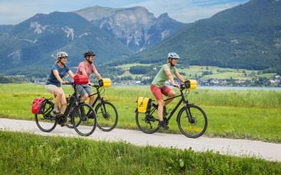 Drie fietsers met fietstassen op een pad langs het Wolfgangsee in Salzkammergut, met bergen en een dorp op de achtergrond.