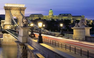 Verlichte Kettingbrug van Boedapest bij schemering met stenen leeuwenbeelden over de Donau. Koninklijk Paleis met groene koepel op de heuvel.