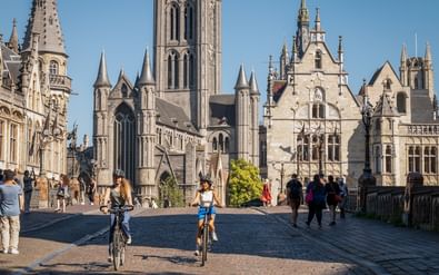 Twee fietsers op geplaveide plein in Gent met Sint-Niklaaskerk en historische gebouwen. Voetgangers onder heldere blauwe lucht.