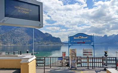 St. Wolfgang Strobl bootaanlegsteiger aan Wolfgangsee met blauw bord, welkomstscherm en bergmeer achtergrond onder bewolkte hemel.