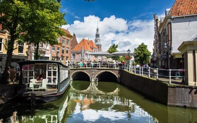 Historische grachtenrug in Alkmaar Stenen brug over gracht in Alkmaar met traditionele Nederlandse gebouwen, kerktoren en toeristenboot. Blauwe lucht met witte wolken weerspiegelt in water.