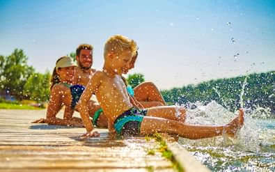 Gezin met twee kinderen spetterend op een houten steiger aan het Brombachsee, met heldere blauwe lucht en groene bomen op de achtergrond.