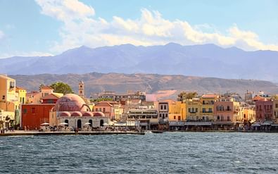 Waterkant van Chania haven met kleurrijke mediterrane gebouwen, waaronder een opvallende koepelmoskee, tegen bergen onder bewolkte hemel.