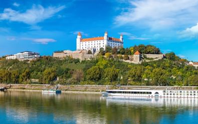 Wit riviercruiseschip op de Donau met kasteel Bratislava op beboste heuvel. Het kasteel heeft witte muren en rode daken onder blauwe lucht.