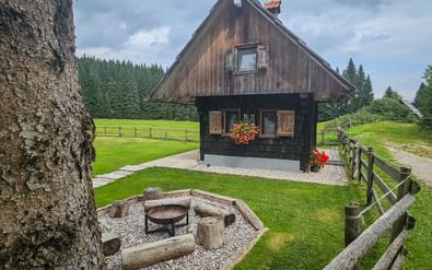 Donkere houten hut met steil dak in Pokljuka, met vuurplaats, groene weiden, houten hekken en bos op de achtergrond.