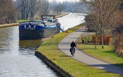 Fietser rijdt over verharde pad langs kanaal in Elzas met afgemeerde vrachtschepen en kale winterbomen langs de waterweg.