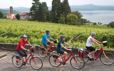 Vier fietsers met helmen op rode fietsen pauzeren op een weg in Piëmont, met wijngaarden, meer en bergen op de achtergrond.