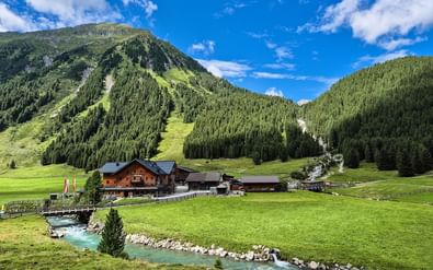 Krimmler Tauernhaus berghut met traditionele houten gebouwen in groene alpenvallei, turquoise beek stroomt door weilanden.