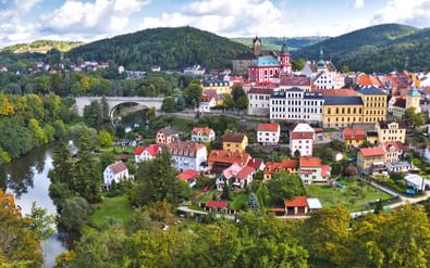 Panoramisch uitzicht op Loket, Tsjechië, met kleurrijke gebouwen, rode kerktoren, stenen brug over de Ohře, omringd door groene heuvels.