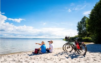Twee fietsers zitten op een zandstrand met verrekijker, wijzend naar de zee. Twee toerfietsen met tassen staan nabij onder een blauwe lucht.