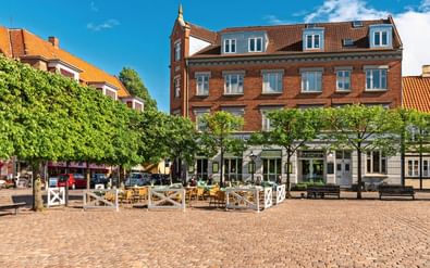 Geplaveide plein in Odense met café-terras, rode bakstenen gebouwen en groene bomen onder blauwe lucht.