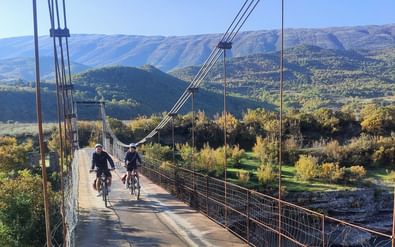 Twee fietsers rijden over een hangbrug met berglandschap in Zuid-Albanië. Groene heuvels en valleien zichtbaar op de achtergrond.