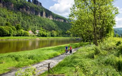 Twee fietsers op verharde weg bij Bad Schandau naast kalme rivier, met dramatische zandstenen kliffen en groen bos op achtergrond.