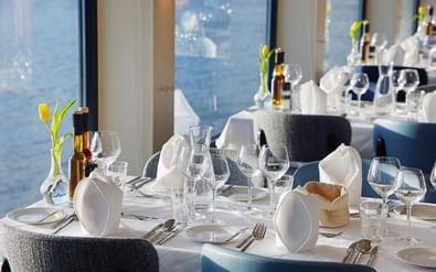 Elegante eetzaal op MS Lisabelle met witte tafelkleden, wijnglazen en blauwe stoelen. Grote ramen tonen zeezicht met gele bloemen.