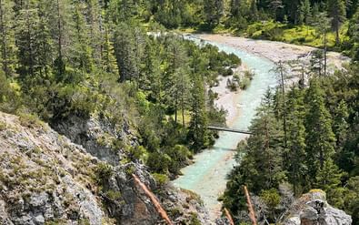 Turquoise Isar-rivier stroomt door rotsachtige bedding omgeven door dicht bos. Een kleine voetbrug kruist de rivier bij de Münchner merenregio.