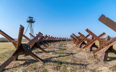 Rijen roestige metalen tankversperringen bij het IJzeren Gordijn Monument in Šatov, Tsjechië, met een wachttoren op de achtergrond onder blauwe lucht.