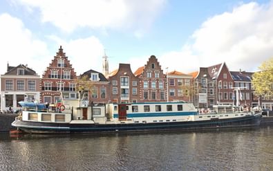Groot wit-blauw historisch schip Flora aangemeerd langs een gracht met traditionele Nederlandse panden en een kerktoren.