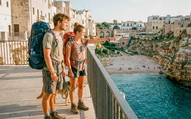 Twee wandelaars met rugzakken op een promenade in Polignano a Mare, Apulië, met uitzicht op een klein strand tussen witte gebouwen en rotsen.