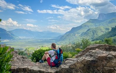 Wandelaar met blauwe rugzak zittend op rotsuitstulping met uitzicht op groene vallei in Zuid-Tirol met bergen en blauwe lucht.