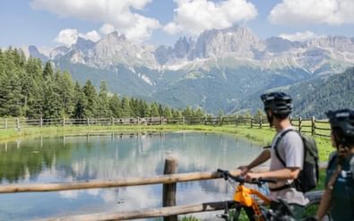 Twee fietsers met rugzakken pauzeren bij een houten hek aan een rustig bergmeer in de Dolomieten, met dramatische rotspieken en bossen op de achtergrond.