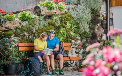 Twee wandelaars zitten op een houten bank en bestuderen een kaart, omringd door kleurrijke bloemen en planten op een stenen muur in de Alpe-Adria-regio.