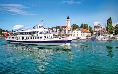 Witte passagiersveerboot Thurgau varend op turquoise water van Bodenmeer met haven van Romanshorn, kerktoren en jachthaven op achtergrond.