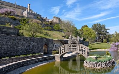 Stenen brug met balustrades over een vijver bij kasteel Stanjel, Slovenië. Historische stenen gebouwen op heuvel met terrasmuren en lentegroen.