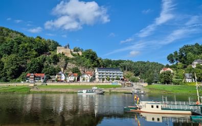 Rivierdorp met boten aan rustig water, huizen langs de oever en een kasteelruïne op een beboste heuvel onder blauwe lucht.