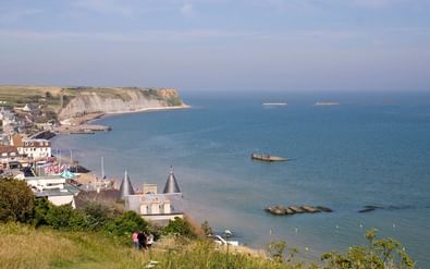 Luchtfoto van een Normandische kustplaats met witte krijtrotsen, blauwe zee, historische gebouwen en D-Day landingsstructuren.
