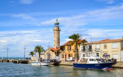 Stenen vuurtoren in de haven van Le Grau-du-Roi met vissersboten, mediterrane huizen, palmbomen en blauwe lucht in de Camargue, Provence.