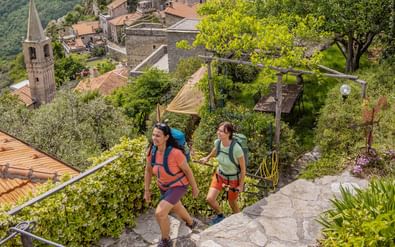 Twee vrouwelijke wandelaars met rugzakken lopen op stenen treden in Castelvecchio, met historische dorpsgebouwen en klokkentoren op achtergrond.