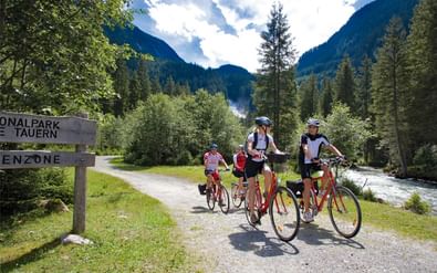 Familie fietsers met rode fietsen bij het ingangsbord van Nationaal Park Hohe Tauern, omringd door bergen, bos en stromende rivier.